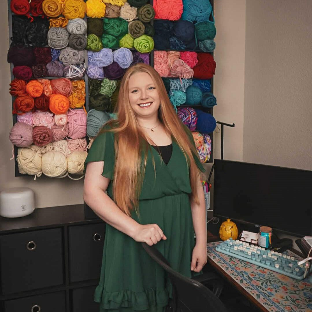 Woman in a green dress standing in front of a colorful yarn wall display.
